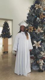 Full length of smiling young woman wearing white graduation gown while standing by christmas tree on hardwood floor