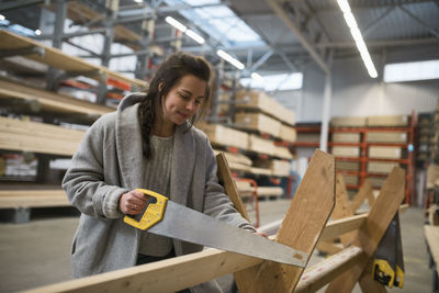 Female customer cutting wooden plank with handsaw in hardware store