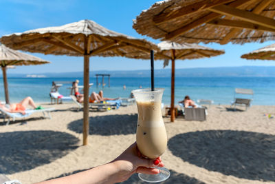 Personal perspective of person holding glass of cold iced coffee on beach in summer.