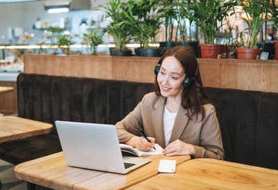 Adult smiling brunette woman forty years in headphones doing notes in daily book with opened laptop