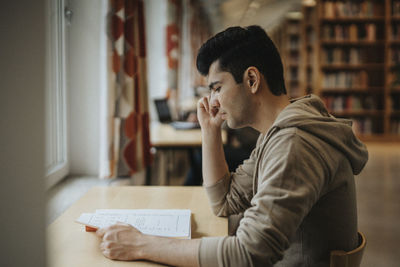 Side view of male student reading book at table in library