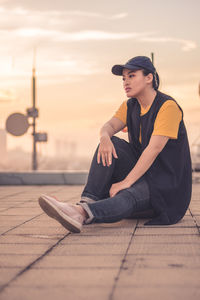 Young woman looking away while sitting on floor against sky during sunset