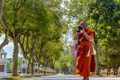 Monk photographing while standing on footpath against trees 