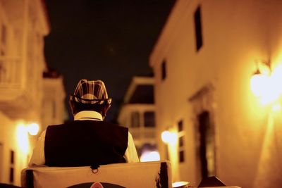 Rear view of man sitting in chair amidst illuminated buildings at night