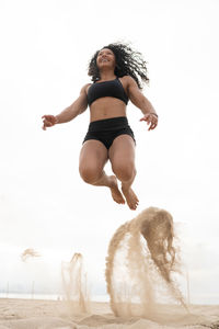 Low angle of cheerful asian female athlete in moment of jumping above sandy seashore during fitness workout in summer