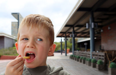 The blond boy looks away and puts french fries in his mouth. a boy has lunch in a street cafe.