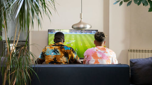 Back view of anonymous multiracial male friends sitting on sofa while playing football videogame on tv in living room with plants