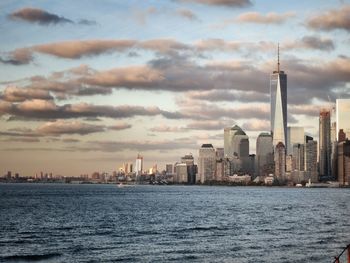 One world trade center and buildings by river against cloudy sky