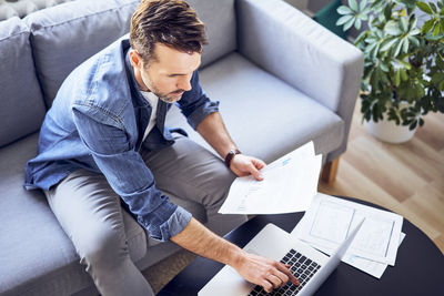 Man with documents sitting on sofa using laptop