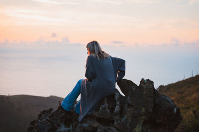 Full body side view of female traveler sitting on stony formation on coast near sea in coastal area on summer evening