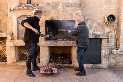 Two adult men are cooking together in the barbecue with more raw meat waiting outside