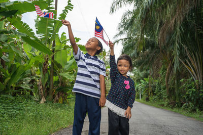 Siblings holding malaysian flag while standing on road amidst plants