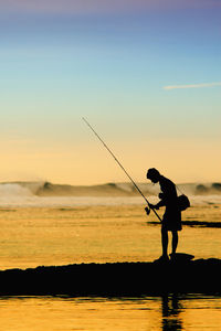 Silhouette man fishing in sea against sky during sunset