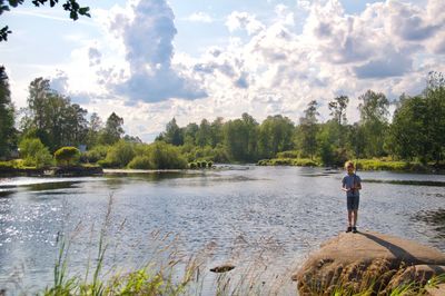 Boy fishing while standing on rock by lake against sky