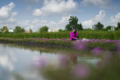 Woman standing by purple flowers on field by lake against sky