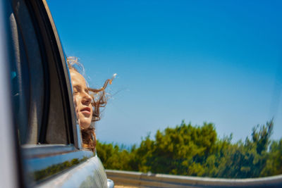 Girl feeling wind through car window against clear blue sky