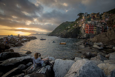 Rear view of woman sitting on rocky shore against cloudy sky