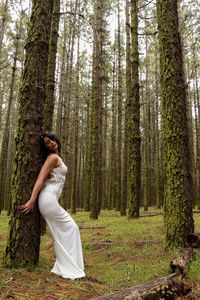 Side view of woman wearing white dress standing by tree trunk in forest