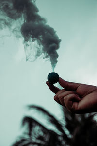 Cropped hand of person holding object emitting smoke against sky