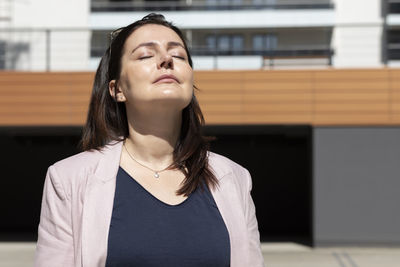 Relaxed mature woman with closed eyes enjoys sun, peace in apartment campus, complex in daytime
