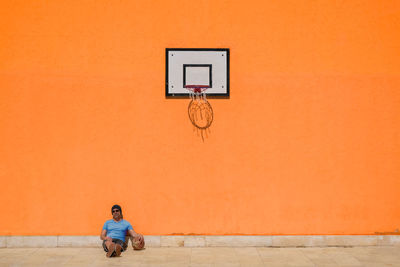 Man sitting on basketball hoop against orange wall