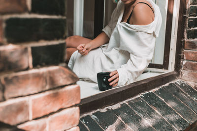 Big black coffee cup in woman's hands with pastel manicure while sitting at windowsill. lifestyle.