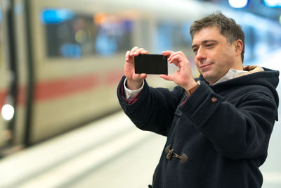 Smiling man photographing with phone at railroad station platform