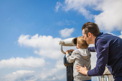 Father and son looking through coin-operated binoculars while standing against sky