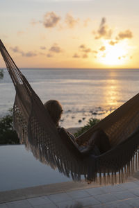 Woman relaxing in hammock at beach against sky during sunset