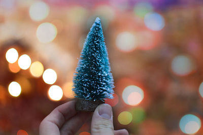 Close-up of human hand holding small christmas tree against illuminated lights