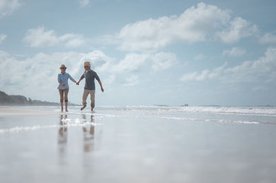 Couple holding hands while walking at beach against sky