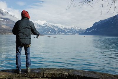 Rear view of person fishing at sea shore against snowcapped mountains