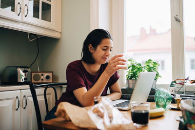 Woman using laptop while having drink at dining table in kitchen