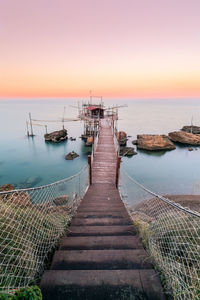 Footbridge leading towards sea against sky during sunset