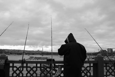 Rear view of man with fishing rods on railing by river against cloudy sky