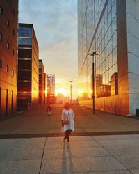 Woman standing on street amidst buildings in city during sunset