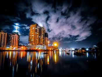 Illuminated buildings by river against sky at night