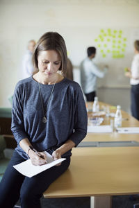 Businesswoman writing on document at conference room with colleagues discussing in background