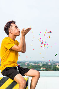 Young man blowing confetti against clear sky