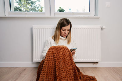 Worried woman sit near heating radiator under blanket and using smartphone