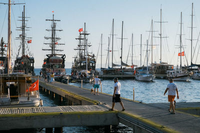 People walking on pier while sailboats moored at harbor against clear sky