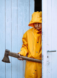 A person in a yellow raincoat and a rusty ax in his hands is hiding behind the ajar door of the barn