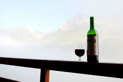 Wineglass and bottle on railing against mountain during foggy weather