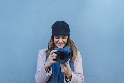 Smiling woman photographing while standing against blue wall