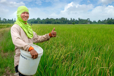 Portrait of smiling farmer gesturing thumbs up while holding bucket on agricultural field