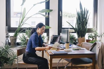 Side view of mature female architect examining documents while sitting at desk in home office