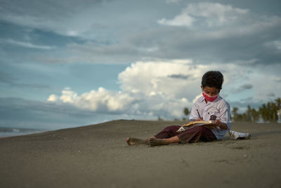 Rear view of boy sitting on sand at beach against sky