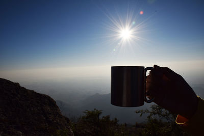 Cropped hand holding mug against sky during sunny day