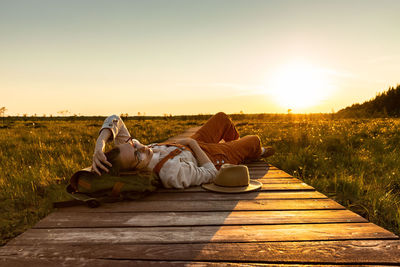 Woman naturalist resting lying on backpack on wooden path through peat bog swamp in wildlife park.