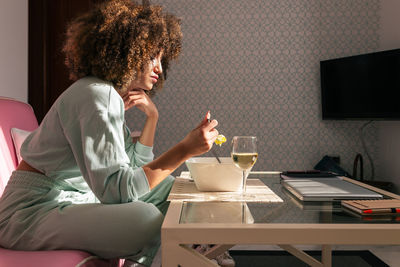 Side view of african american female eating salad while sitting at table with glass of wine and having tasty lunch at home